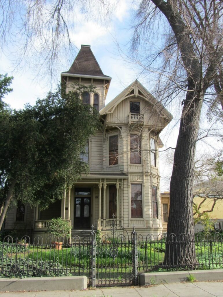 A large, Victorian-style house with intricate architectural details, surrounded by trees and a wrought-iron fence.