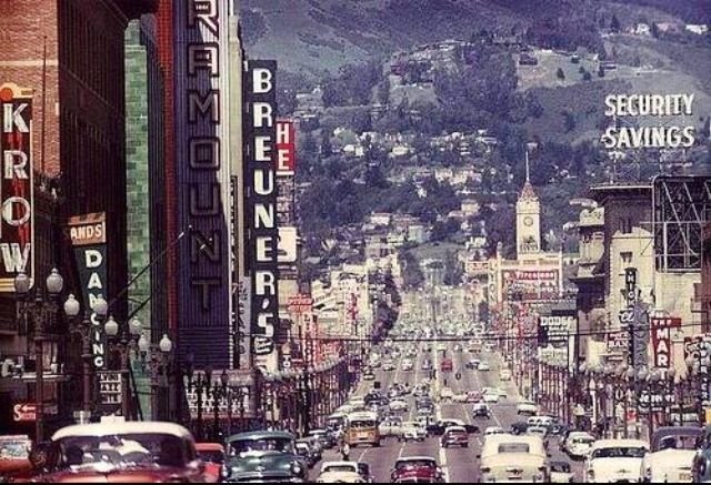 A colorful vintage photo of a busy street lined with shops and cars, showcasing iconic signs from the past.